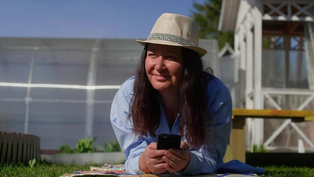 Portrait of dreaming relaxed woman in sun hat with smartphone in hands enjoying resting lying on grass outdoors and looking at blue clear sky. Weekend in summer house, dacha at sunny weather.