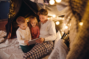 Obraz na płótnie Canvas Young father and his kids watching cartoons on tablet while sitting in tent in children's room