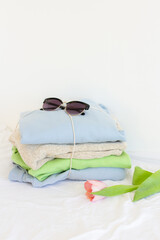 Pile of clothes, sunglasses and one pink tulip on white, pastel color palette of stacked sweaters and jeans, fresh clothing folded for ironing