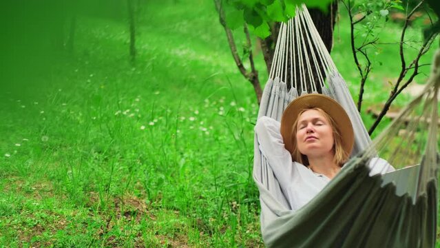 Relaxed female freelancer wearing hat chilling in a hammock among green trees. Charming blonde napping outdoors. Happy young woman enjoying summer vacation.
