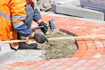 A worker using a rubber mallet and a wooden level lays orange paving slabs evenly on a sunny summer day.
