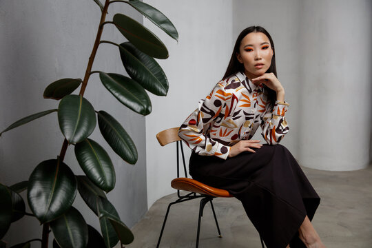 High fashion photo of beautiful elegant young asian woman in pretty beige shirt with pattern, long black skirt posing on textured gray wall, tall flower. Slim figure. Model is sitting on a chair. 