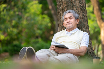portrait healthy asian senior man closed eyes,siting against a tree on grasses while reading and relaxing in the park,concept of old people lifestyle,health benefits in  nature,wellbeing