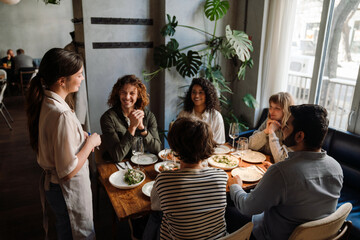 Beautiful waitress serving group of cheerful friends in restaurant