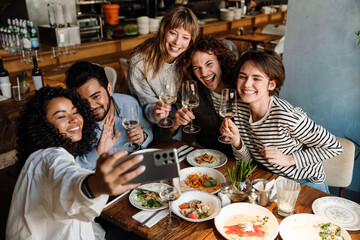 Group of cheerful friends taking selfie and drinking wine while dining in restaurant