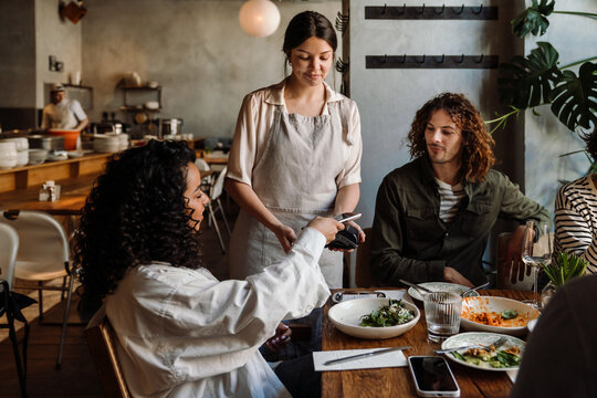 Woman using smartphone while paying restaurant bill with contactless payment during dinner party with friends