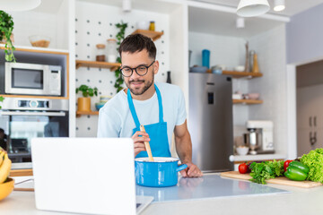 A young man spends a day at home, prepares food of vegetables in the kitchen, a man in home clothes and with a beard, uses a laptop to learn online cooking