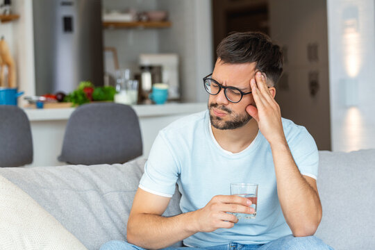 Young man suffering from strong headache or migraine sitting with glass of water on the sofa, millennial guy feeling intoxication and pain touching aching head, morning after hangover concept