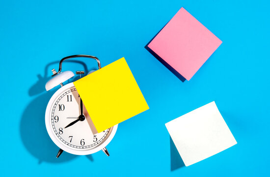 Alarm clock and colorful blank paper reminders on a blue background, top view.