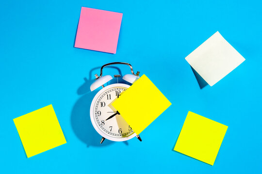 Alarm clock and colorful blank paper reminders on a blue background, top view.
