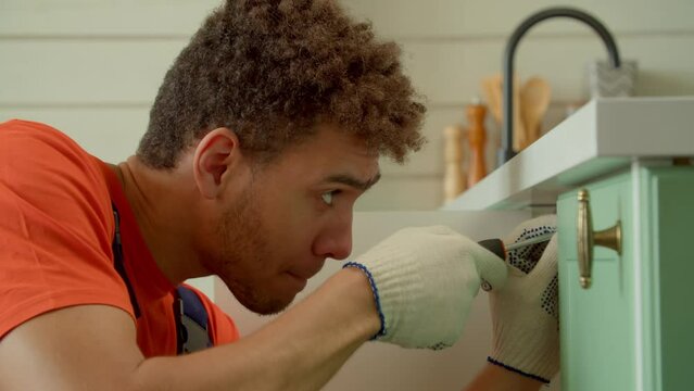 Portrait of concentrated attractive black handyman in protective workwear doing home DIY, repairing broken kitchen cabinet, and fixing tapping screws using screwdriver indoors.