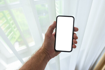 Male hand with mockup smartphone next to the living room window with white curtains on sunny summer day