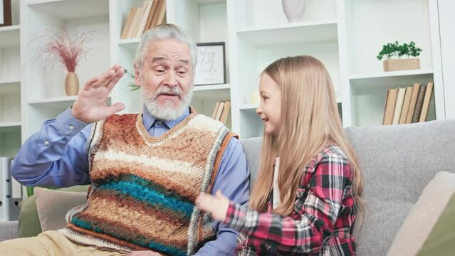 Charismatic grandfather teaching child how to give high five and laughing. Open-minded girl having rest at home and playing with grandfather. Concept of happiness and sincere emotions.