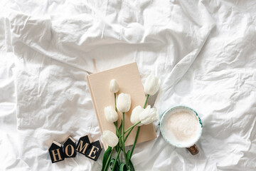 Fototapeta na wymiar A bouquet of white tulips, a cup of coffee and a book in a white bed, top view.