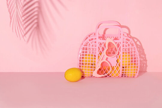 Summer creative layout with pink jelly basket, heart sunglasses with lemons and palm tree shadow on pink background. 80s or 90s retro aesthetic idea. Minimal summer fruit idea.