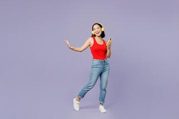 Full body young woman of Asian ethnicity she wears casual clothes red tank shirt headohones listen to music use mobile cell phone isolated on plain pastel light purple background. Lifestyle concept.