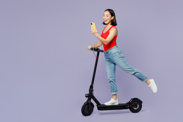 Full body sideways young woman of Asian ethnicity she wear casual clothes red tank shirt riding electric scooter use mobile cell phone isolated on plain pastel light purple background studio portrait