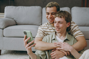 Young fun couple two gay men wear casual clothes together hold use mobile cell phone sit on sofa...