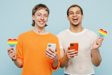 Young smiling couple two gay men wearing casual clothes together use mobile cell phone hold paper hearts isolated on pastel plain blue color background studio. Pride day june month love LGBTQ concept.