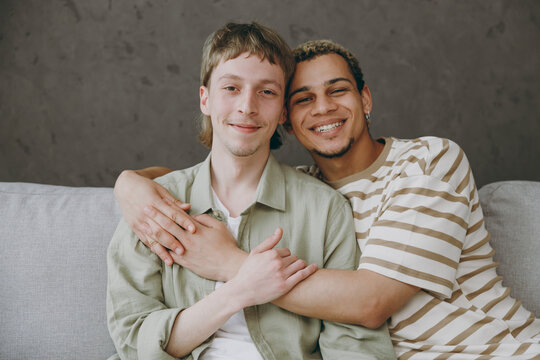 Young happy calm couple two gay men wear casual clothes together hugging look camera sit on sofa couch at home flat rest spend free spare time in living room. Pride day june month love lgbtq concept.