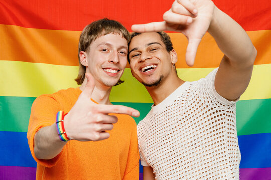 Young happy couple two gay men wear casual clothes together do photo frame with hands pov taking picture isolated on striped flag background studio portrait. Pride day june month love LGBTQ concept.