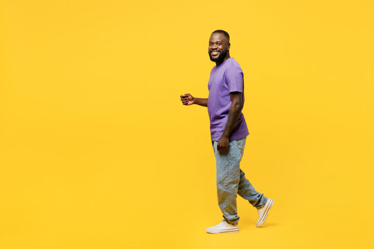 Full Body Side View Young Man Of African American Ethnicity He Wear Casual Clothes Purple T-shirt Looking Camera Walk Go Stroll Isolated On Plain Yellow Background Studio Portrait. Lifestyle Concept.