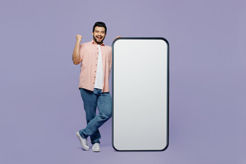 Full body young Indian man he wears pink shirt white t-shirt casual clothes big huge blank screen mobile cell phone smartphone with area do winner gesture isolated on plain pastel purple background.