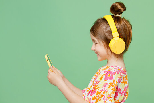 Side view little child kid girl 6-7 years old wear casual clothes headphones listen music use mobile cell phone isolated on plain pastel green background. Mother's Day love family lifestyle concept.