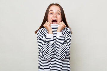 Extremely surprised positive overjoyed woman with brown hair wearing striped shirt standing isolated over gray background screaming with excitement.