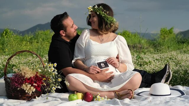 In slow motion, a pregnant woman with a crown of daisies and her partner in black sit on a picnic blanket on the grass, holding their baby's ultrasound images, immersed in serene anticipation