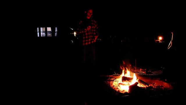 Man having drink while standing next to bonfire in nigh with only flames as light source illuminating his body and face and some lights visible in the background from mobile house windows.