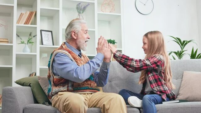 Caring grandfather spending weekend with little granddaughter and having fun together. Happy child and grandfather playing hand-clapping game while resting on soft couch. Concept of family bond.