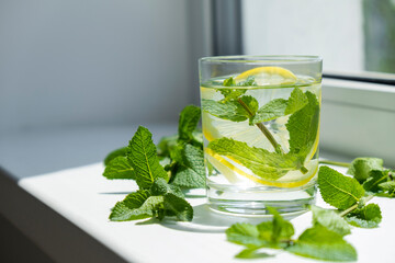 Glass with cold lemonade or mojito on window sill and fresh mint leaves around. Herbal refreshing drink. Homemade antioxidant water.
