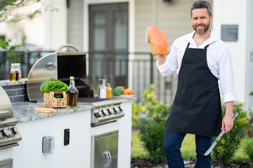 Man in chef apron hold salmon fish at barbecue grill. Male cook preparing barbecue outdoors. Bbq fish, grill for picnic. Roasted salmon fish. Grilled fish and barbecue seafood concept.