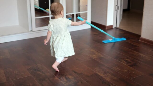 Toddler daughter helps mother to clean spacious room floor using wet mop. Barefoot girl wearing dress enjoys cleaning apartment independently