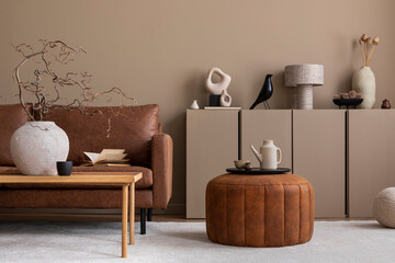 Warm and cozy living room interior with brown sofa, wooden coffee table, round pouf, vase with branch, beige sideboard, pitcher, cup, books and personal accessories. Home decor. Template.