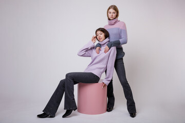 Two fashion models in pastel lilac and pink sweater, blouse, black jeans, denim. Beautiful young woman. Studio shot, portrait. White background. Asian and  blonde. Slim figure. Model sits on cube