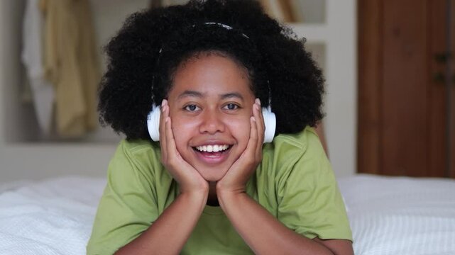 Attractive curly-haired black teen girl with headphones on bed laughs and smiles with her teeth while looking at camera. Multinational multiracial teen girl laughs and smiles, close-up, portrait shot