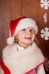 A girl in a warm red scarf and a santa claus hat examines paper snowflakes. The child rejoices in winter, New Year cartoons and the holiday of Christmas