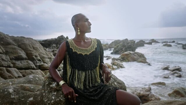 Gay bipoc man in luxury dress, boutique brass jewelry, makeup poses sitting on tropical rocky ocean beach. Homosexual epatage fashion model in posh gown, nose-ring piercing, earrings, rings with gems.