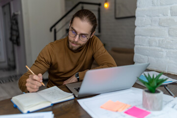 Fototapeta premium Man using laptop writing down important data in notebook, working on computer or studying online.