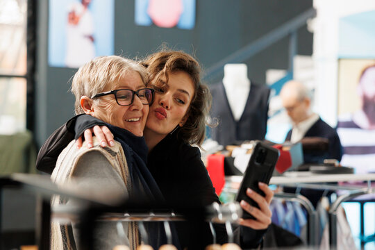 Cheerful women taking selfie with modern phone, smiling at camera in clothing store. Customers shopping for casual wear, buying stylish clothes and fashionable merchandise. Fashion concept