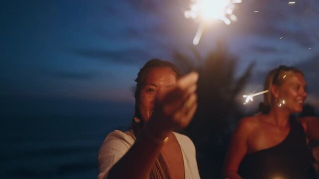 Diverse women dance, laugh with sparklers at seaside tropical hen party at sunset. Young smiling multiracial females have fun at 4th July Independence Day celebration by sea with sparkling lights