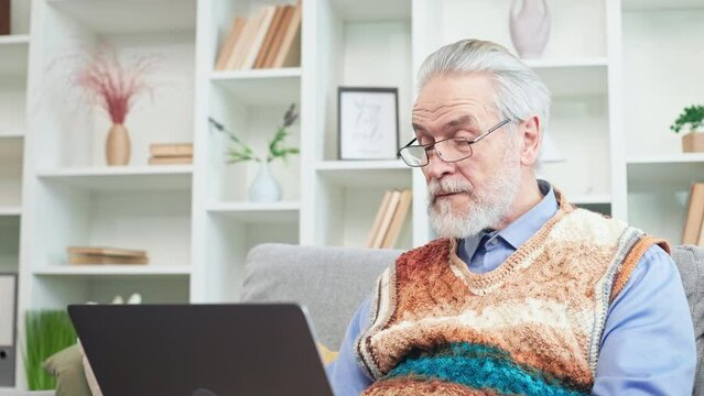 Concentrated man on retirement chatting in social media with family and friends using wireless laptop. Casually dressed progressive gentleman in glasses relaxing on couch and communicating online.