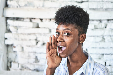 A miggle-aged african woman sitting on her sofa shouting and holding palm near opened mouth.