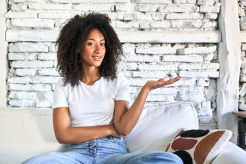 A young african american woman sitting on her sofa showing a copy space on a palm and holding...