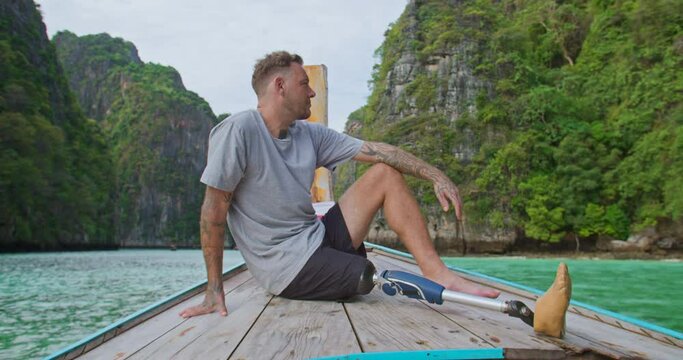Young caucasian male with artificial bionic legs prosthesis sitting on the front of long tail boat seeing an amazing beautiful seascape during summer vacation travel in Phi Phi Islands, Thailand.