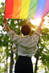 Rear view of young woman holding and raising a rainbow flag with sunlight shining through green...