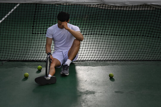 Disappointed asian male tennis player sitting on the court, upset about competition loss, failure emotions