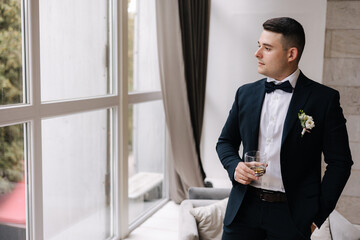 Young groom stand near big window and hold glass of whiskey at home. Morning of groom at wedding day